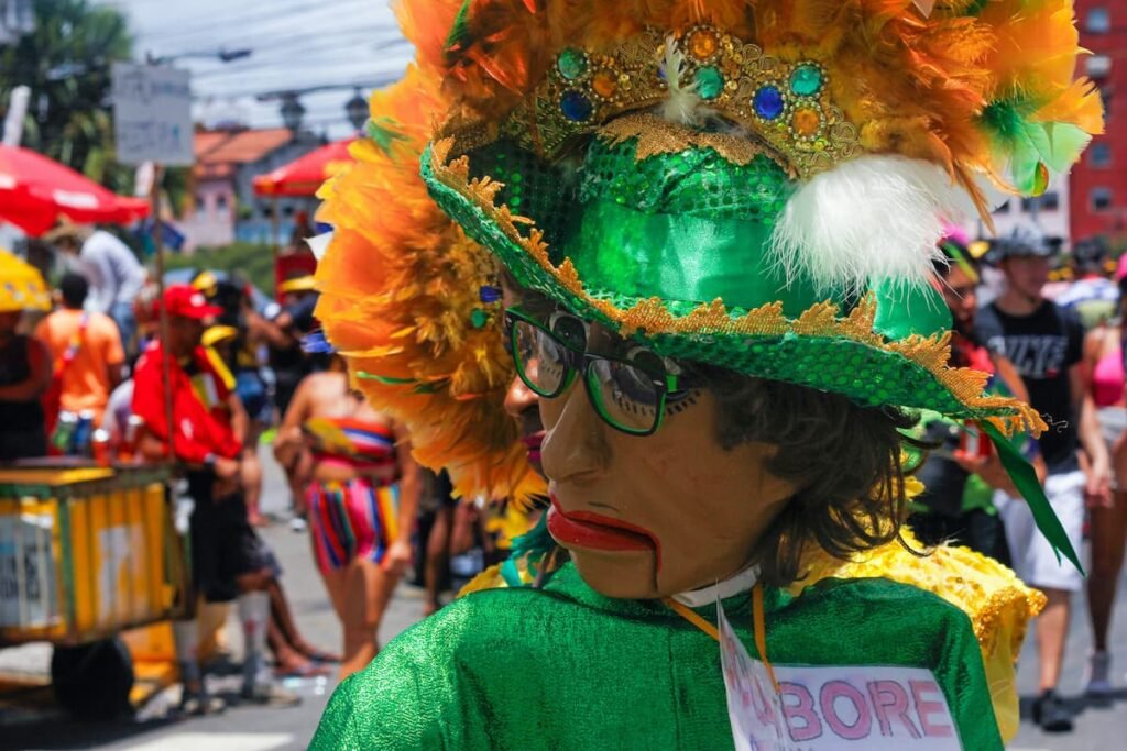 Boneco em bloco de rua no carnaval do Rio de Janeiro, usando traje vibrante com penas laranja e amarelas, máscara expressiva com óculos, e roupa verde e dourada. Ao fundo, foliões celebram em clima festivo e colorido.