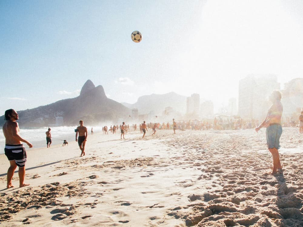 Praia de Ipanema em dia ensolarado, com pessoas jogando futebol na areia e a bola no ar.