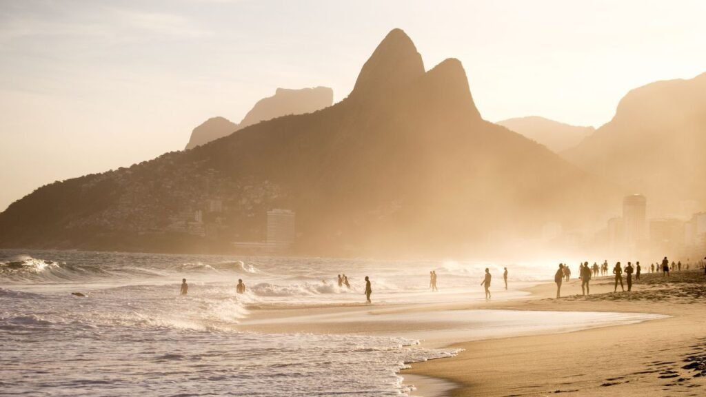 Praia de Ipanema ao pôr do sol, com pessoas caminhando na areia e nadando no mar.