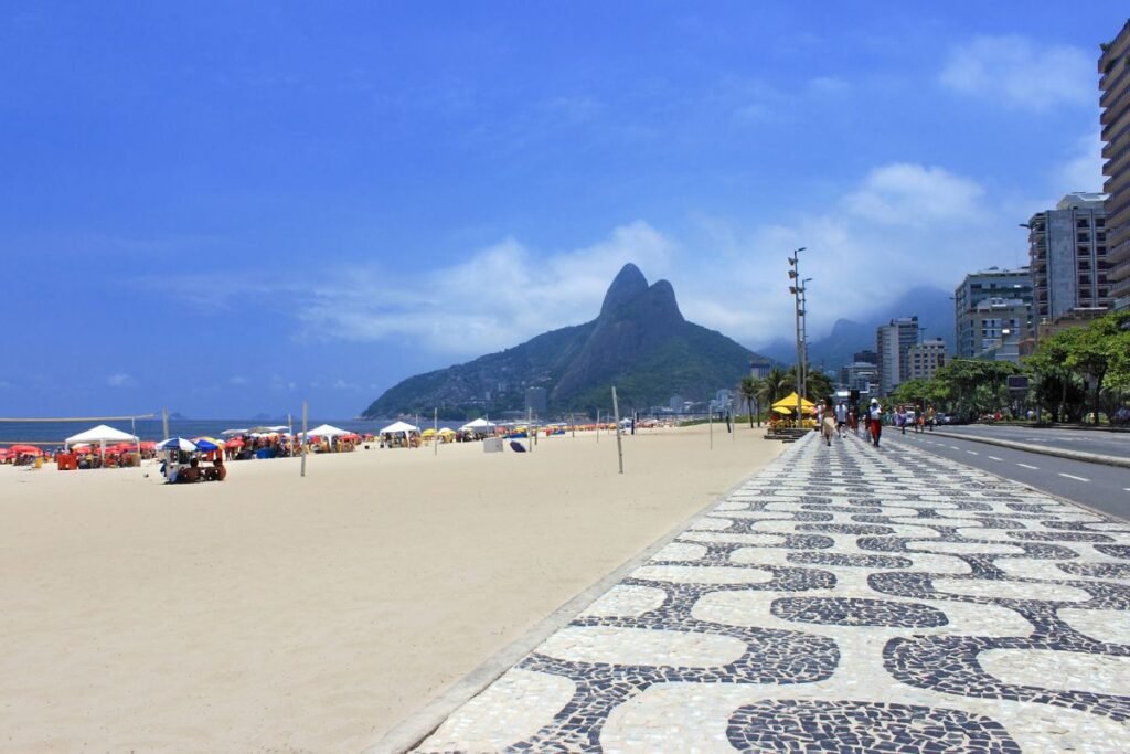 Vista vibrante da Praia de Ipanema, com o calçadão em mosaico preto e branco em primeiro plano, areia repleta de guarda-sóis coloridos e o Morro Dois Irmãos ao fundo envolto em névoa.