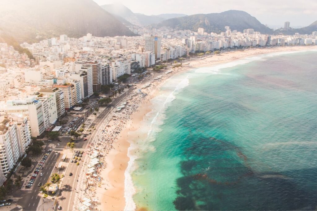 Vista aérea da Praia de Copacabana, mostrando a extensa faixa de areia cheia de banhistas, mar azul-turquesa e a avenida costeira ladeada por prédios altos com montanhas verdes ao fundo.